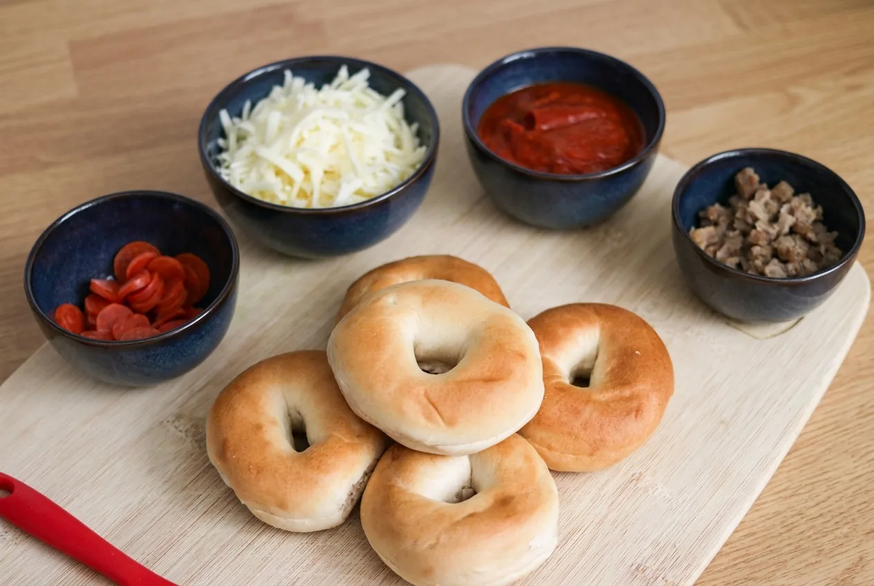 Félbevágott mini bagelek deszkán, mellettük reszelt sajt, pizzaszósz, pepperoni és kolbász kék tálkákban
