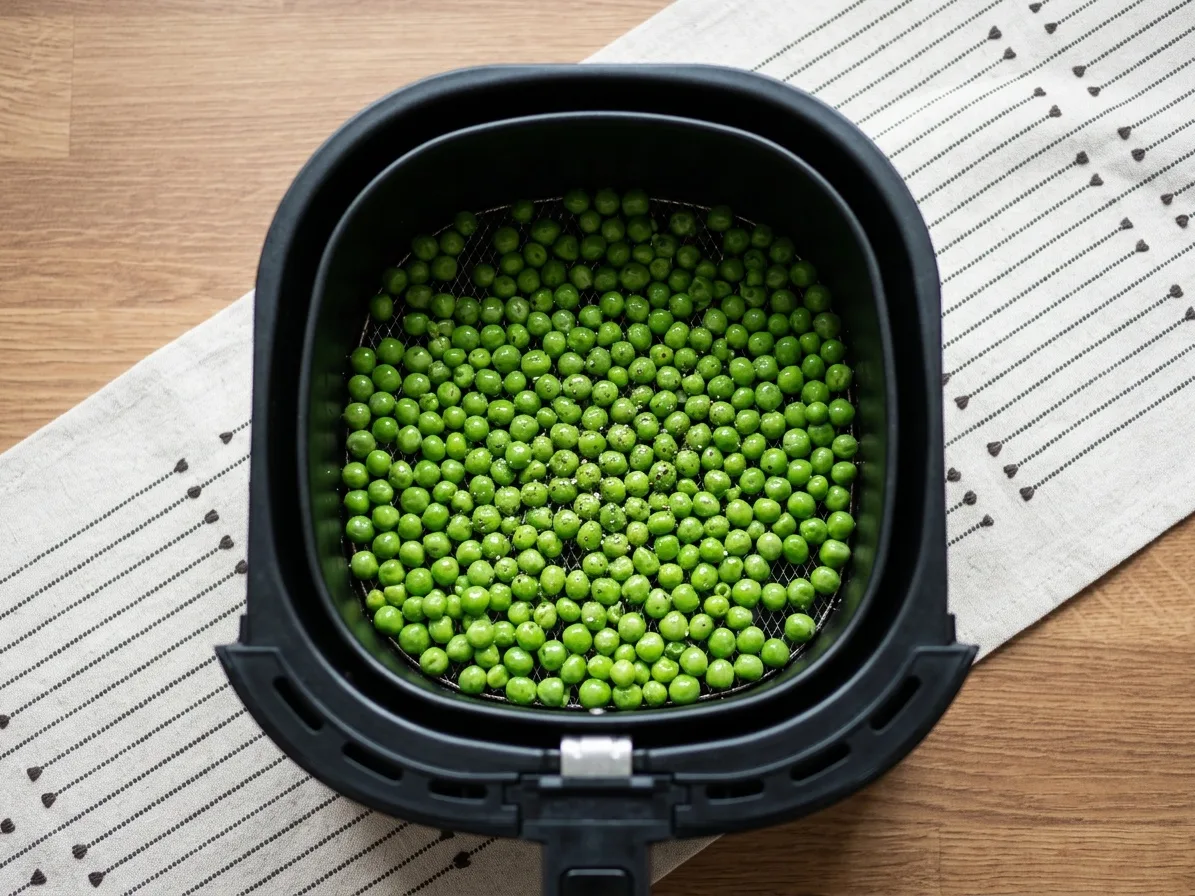Olajjal, sóval és borssal összeforgatott zöldborsó egy rétegben az air fryer kosarában, sütés előtt